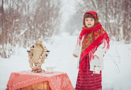 Adorable little girl in old Russian clothes in a winter forest with a samovar and bagelsの写真素材