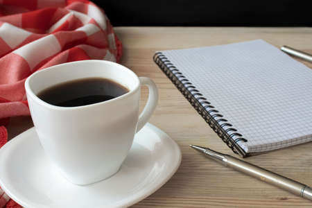 White cup of strong black coffee with red-white cloth, metal pen and notepad on wooden surface close upの写真素材