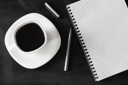 White cup of strong black coffee with metal pen and notepad on black surface close upの写真素材