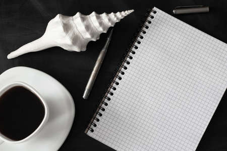 White cup of strong black coffee with metal pen, notepad and seashell on black surface close upの写真素材