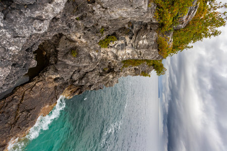 Limestone Wall Rugged Rocky Cliff with Trees Surrounded by Turquoise Water of Lake Huron, vertical shot during dramatic stormy autumn day at Indian Head Trail, Ontario, Canadaの写真素材