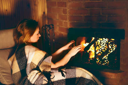 a red-haired beauty wrapped in a blanket is warming herself near the fireplace and drinking tea.の写真素材