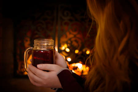 A young girl with long hair is sitting in front of the fireplace, holding a cup of tea and warming herself. the concept of warmth, dreams and happiness. Close-up, selective focusの写真素材