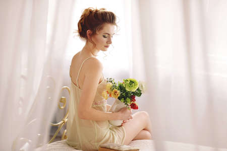 Beautiful young woman in a sexy nightgown. morning dreams of a girl in love. The bouquet in your hands reminds you of your beloved man. a room for a girl. playful mood. about loveの写真素材
