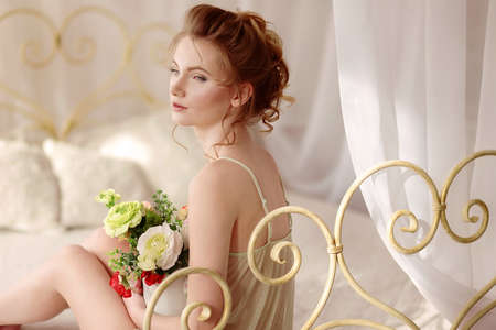 Beautiful young woman in a sexy nightgown. morning dreams of a girl in love. The bouquet in your hands reminds you of your beloved man. a room for a girl. playful mood. about loveの写真素材