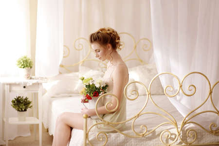 Beautiful young woman in a sexy nightgown. morning dreams of a girl in love. The bouquet in your hands reminds you of your beloved man. a room for a girl. playful mood. about loveの写真素材