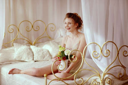 Beautiful young woman in a sexy nightgown. morning dreams of a girl in love. The bouquet in your hands reminds you of your beloved man. a room for a girl. playful mood. about loveの写真素材