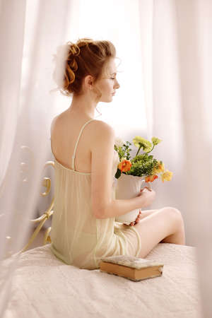 Beautiful young woman in a sexy nightgown. morning dreams of a girl in love. The bouquet in your hands reminds you of your beloved man. a room for a girl. playful mood. about loveの写真素材