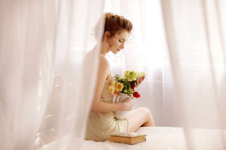Beautiful young woman in a sexy nightgown. morning dreams of a girl in love. The bouquet in your hands reminds you of your beloved man. a room for a girl. playful mood. about loveの写真素材
