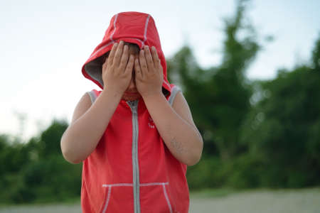 Little adorable boy covers his eyes with his hands standing outdoors. he is agitated, razdasadavan and wants to cry because he fell and hurt his elbow. closed from pain and resentment. manipulates.の写真素材