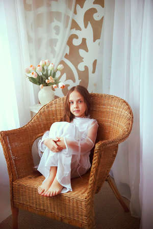 a little girl sits in a wicker chair and dreams of going out in the evening to run with the boys and girls. portrait of a young lady. the concept of a happy family and a carefree childhood.の写真素材