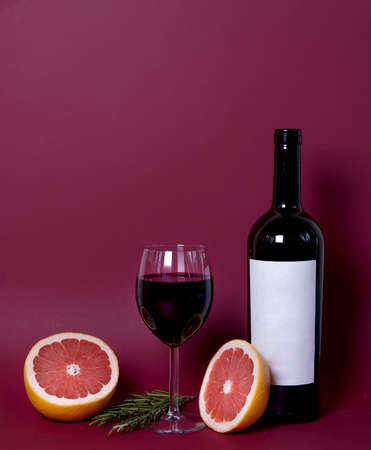 a glass of red wine on a burgundy background. red grapefruit and rosemary sprigs give the composition a special unique style. copy space. advertising a restaurant, winery, or wine list.の写真素材