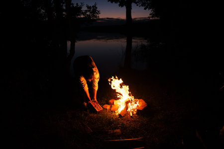 The silhouette of a girl by the fire on the lake shore. night in the forest. night shooting, soft focus. advertising of tourism companies and fishing accessories. the concept of recreationの写真素材