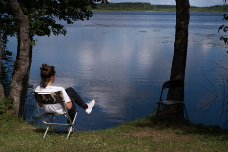 A tourist girl is resting, drinking tea on the shore of the lake. the girl is enjoying her summer vacation. The concept of a quiet and peaceful country holiday away from civilization.の写真素材