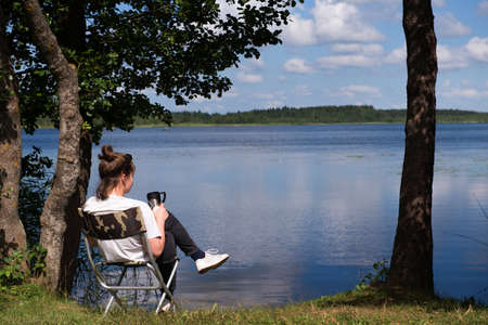 A girl tourist-traveler is resting, drinking tea on the shore of the lake. the girl is enjoying a summer vacation. The concept of a quiet and peaceful country holiday away from civilization.の写真素材