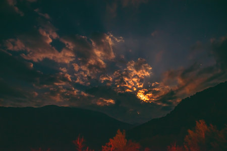 The orange full moon rising from behind the mountain range illuminates the misty clouds, creating a...