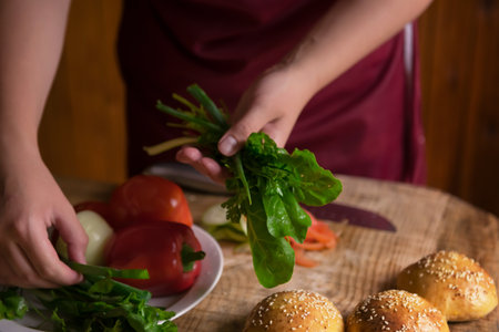 A young woman cuts fresh vegetables on a wooden salad board. Cooking. A girl cuts vegetables in close-up. cooking a hamburger at home. vegetarianism. selective focus, photo in a dark keyの写真素材