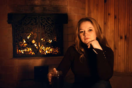 A beautiful young woman is sitting by the fireplace in a dark room at home and drinking a hot drink from a large cup. A beautiful girl enjoys tea, coffee in a cozy room. Christmas scene. retro styleの写真素材