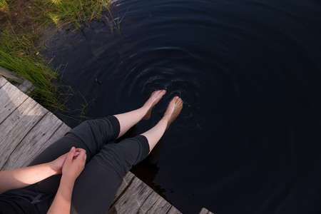 A girl is resting by the lake, sitting on the edge of a wooden pier, her legs dangling along the surface of the water. Tired of the summer heat, she cools down by the water. Hot summer day conceptの写真素材