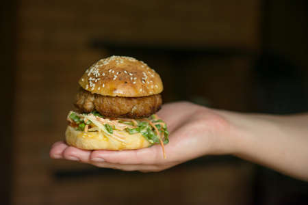 a woman's hand holds a hamburger on a dark background. Cooking a hamburger at home, home cooking. fast food at home, special offer. author's cheeseburger recipes. selective focus, copy spaceの写真素材