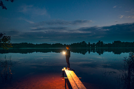 a young girl with a flashlight stands on the shore of a forest lake, as the lighthouse illuminates the way to the shore. Waiting for a guy from fishing. evening comes, the first stars appear in the skyの写真素材