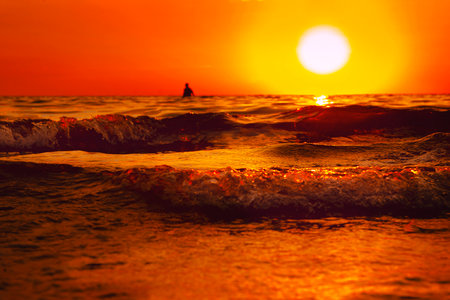 The sun is sinking into the sea. Bright orange rays pass through the waves, rolling smoothly. In the...