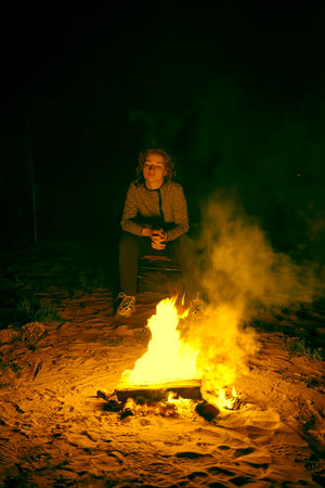 A girl is sitting at night by a campfire on the seashore, a small fire is lit on the sandy shore. the logs are burning down, and the girl is warming herself by the fire. the concept of rest and travelの写真素材