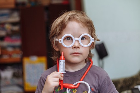 A little boy is playing doctor. Little Caucasian boy doctor. A shot, a syringe in his hand. children playing doctor and curing plush toy at homeの写真素材