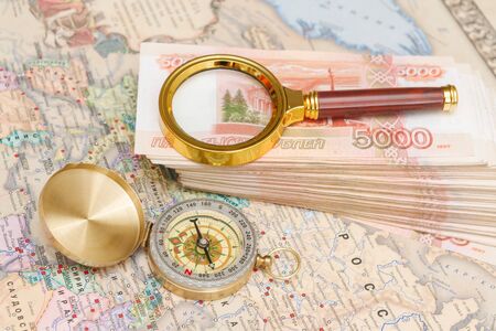 A compass with a bundle of big money and a magnifying glass are on the map