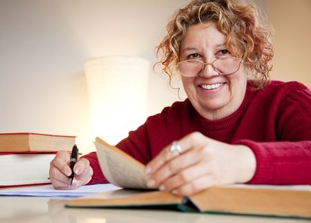 mature female is sitting by a table with books and a pen, smilingの写真素材