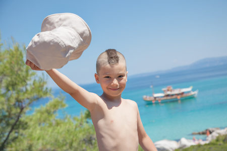 A young boy is waving with his hat by a beautiful seashore with departing ship.の写真素材