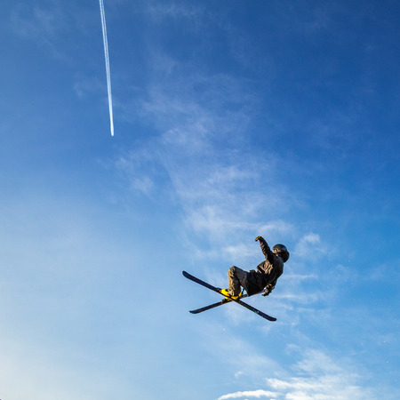 Ski jumper flying high in the air on a blue sky background with a trace of a jet aircraft.の写真素材