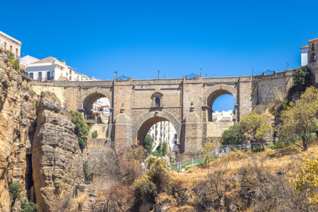 The Puente Nuevo bridge in El Tajo gorge in Ronda town in Spain. Ancient stone bridge spanning a deep gorge, with buildings visible on either side under a clear blue sky.の写真素材