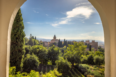 Alhambra complex in Granada town, Andalusia, Spain. Scenic view of a historic city from a garden archway. Lush greenery and distant buildings under a bright sky.の写真素材