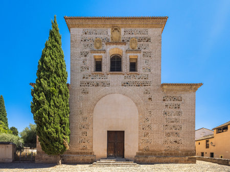 Church of Santa Maria at the Alhambra complex in Granada town, Andalusia, Spain. Ancient stone building with arched entryway, under a clear blue sky. A tall, slender evergreen tree stands beside it.の写真素材