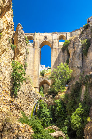 The Puente Nuevo bridge in El Tajo gorge in Ronda town in Spain. Ancient stone bridge spanning a deep gorge, lush vegetation clinging to the rocky walls. A small waterfall cascades down.の写真素材