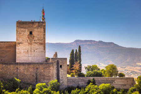 View of the Alhambra palace from the Mirador de San Nicolas lookout in Granada town in Spain. Ancient stone tower overlooking a mountain range. People stand on the tower's top, enjoying the view.の写真素材