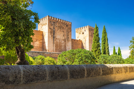 The Alcazaba fortress at the Alhambra complex in Granada town, Andalusia, Spain. Ancient stone towers stand tall against a vibrant blue sky, surrounded by lush greenery.の写真素材