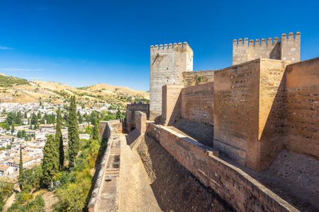 The Alcazaba fortress at the Alhambra complex in Granada town, Andalusia, Spain. Ancient fortress walls overlooking a city nestled in hills. A sunny day reveals the aged stone and distant cityscape.の写真素材