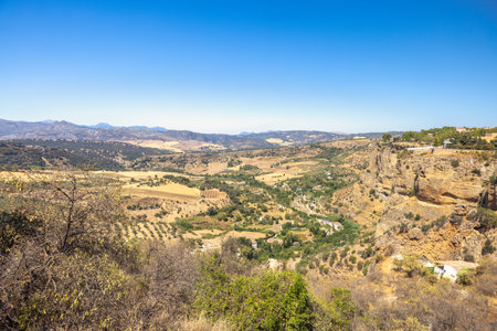 The mountainous land of Andalusia near Ronda town in Spain. Vast valley landscape viewed from a high vantage point, showcasing diverse terrain, vegetation and distant mountains under a clear blue sky.の写真素材