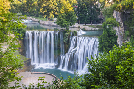 The Pliva Waterfall in Jajce town, Bosnia and Herzegovina, Europe. Majestic waterfall cascades down rocks into a pool, framed by lush green trees, creating a serene natural landscape.の写真素材