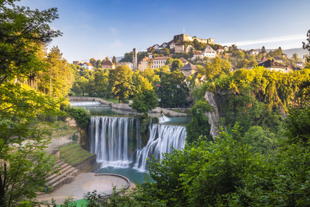 The Pliva Waterfall in Jajce town, Bosnia and Herzegovina, Europe. Picturesque waterfalls cascade in a lush, green setting below a historic town perched on a hillside under a vibrant blue sky.の写真素材