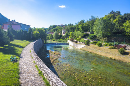 Jajce town in Bosnia and Herzegovina, Europe. Scenic river walk with stone path and clear water, lush greenery, houses dotting the hillside under a bright blue sky. Peaceful outdoor scene.の写真素材