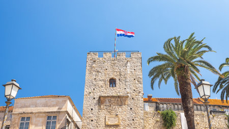 Korcula town in island of Korcula in Adriatic sea in Croatia, Europe. A historical stone tower stands tall against a clear blue sky, topped with a national flag and framed by palm trees.の写真素材