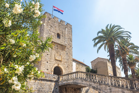 Korcula town in island of Korcula in Adriatic sea in Croatia, Europe. Historic stone tower with a Croatian flag atop, seen through lush greenery and a bridge, under a clear blue sky.の写真素材