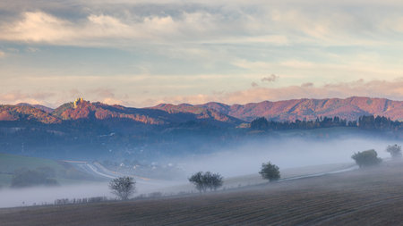 Misty valley landscape with hills, trees, and distant mountains under a pastel sunrise sky.の写真素材