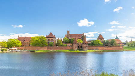 The Malbork Castle, a brick Gothic castle in northern Poland, Europe. Majestic castle complex reflecting in the still waters, embraced by lush greenery under a clear sky with scattered clouds.の写真素材