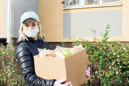 female volunteer wearing a protective mask walks down the street with a box of groceries, donations.の写真素材