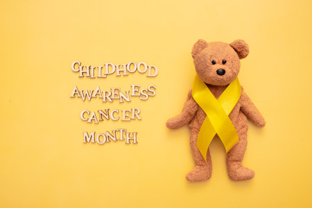 Childhood cancer awareness month text from wooden letters and teddy bear with yellow ribbon on yellow background.の写真素材