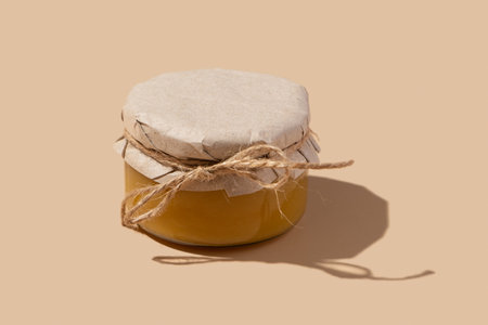 A glass jar with honey under kraft paper on a string on beige background. Sweet food monochrome conceptの写真素材